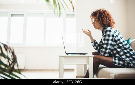 Eine reizende kaukasische Frau mit lockigen Haaren und einer Brille, die auf dem Laptop bei einer Online-Konferenz etwas erklärt Stockfoto