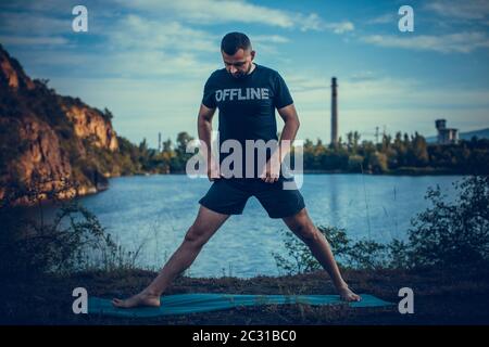 Inspirierter Mann, der Yoga Asanas auf dem Steinbruch-See macht. Junge Bürger trainieren draußen und stehen in Yoga-Seitenwinkel Pose. Fitness im Freien und Life Balanc Stockfoto