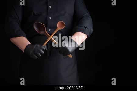 Koch in schwarzen Latexhandschuhen und einer schwarzen Uniform hält gekreuzte Holz-Vintage-Löffel vor sich, Low Key, Copy-Platz Stockfoto