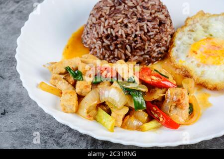 Hähnchen mit süßen Zwiebeln und Paprika unter Rühren braten, serviert mit braunem Reis und Spiegelei auf weißem Teller Stockfoto