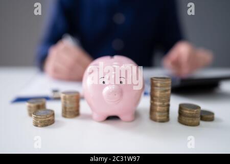 Der Unternehmer Hand mit Taschenrechner mit Gestapelte Münzen und Sparschwein auf hölzernen Schreibtisch Stockfoto