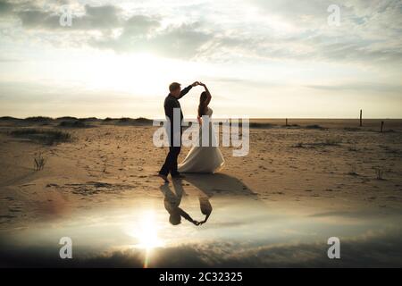 Frisch verheiratetes Paar tanzt am Sandstrand. Braut und Bräutigam am Abend im Hinterlicht. Barfuß tanzen, Spiegelreflexion. Hochzeitskonzept. Stockfoto