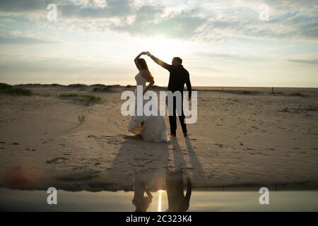 Frisch verheiratetes Paar tanzt am Sandstrand. Braut und Bräutigam am Abend im Hinterlicht. Barfuß tanzen, Spiegelreflexion. Hochzeitskonzept. Stockfoto