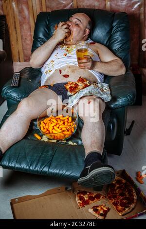 Fat couch potato Essen einer riesigen Hamburger und Fernsehen. Grelle Beleuchtung aus dem Fernsehen beleuchtet die dunklen Zimmer. Stockfoto