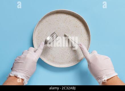 Hände in Handschuhen halten Gabel und Messer auf hellblauem Hintergrund in der Draufsicht über die Platte. Ostern beim Coronavirus Konzept. Stockfoto