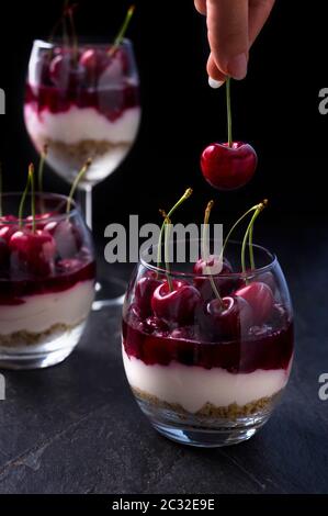 Geschichteter Kirschkäsekuchen in einem Glas. Dessert aus Keksen, Ricotta, Sahne, Kirschgelee und frischen Früchten. Dunkle launische Foto. Stockfoto