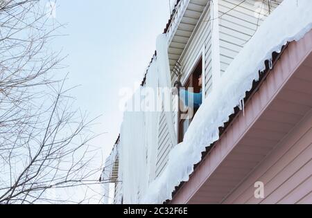 Ein Mann entfernt an einem klaren Wintertag auf dem Land riesige Eiszapfen und Schnee durch das Fenster mit Hilfe einer Mistgabel vom Dach. Saisonales Hauptgebäude Stockfoto