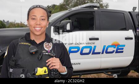 Eine freundliche junge Polizistin gemischter Ethnie lächelt für die Kamera, während sie vor einem Polizeiwagen steht. Stockfoto