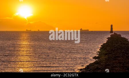 Silhouette von Frachtschiffen. Sonnenscheibe, leuchtet hinter Wolke. Leuchtturm auf Bull Island Stockfoto