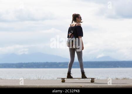 Eine junge Frau reitet an einem sonnigen Nachmittag in Seattle, Washington, auf einem Skateboard entlang des Alki Trail. Stockfoto