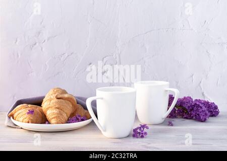 Zwei schöne Porzellanbecher Kaffee mit Milch mit Croissants mit Fliederblumen auf weißem Holztisch dekoriert. Perfektes Frühstückskonzept. Kopieren Stockfoto