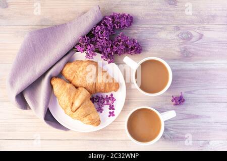 Zwei schöne Porzellanbecher Kaffee mit Milch mit Croissants mit Fliederblumen auf weißem Holztisch dekoriert. Perfektes Frühstückskonzept. Flach l Stockfoto