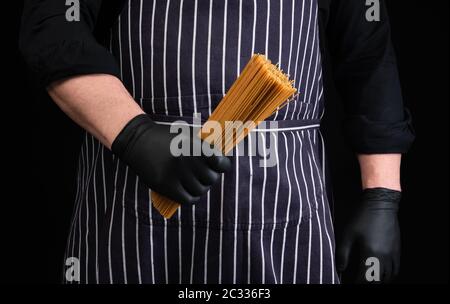 Chefkoch in schwarzen Latexhandschuhen, gestreifte Schürze hält roh lange gelbe Spaghetti aus Hartweizen aus nächster Nähe Stockfoto