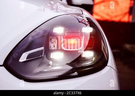 Linke LED-Scheinwerfer eines weißen Premium-suv. Mit rot-orangefarbener Beleuchtung von der Linse. Neue Generation von Autolampen. Nahaufnahme einer Haube und eines Stoßfängers. Stockfoto