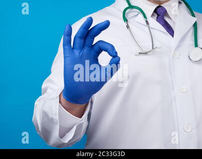 Männliche Ärztin in weißem Mantel, blaue Latexhandschuhe zeigen mit der rechten Hand eine gute Geste, das Konzept der Hoffnung, dass alles in Ordnung sein wird, blauer Backgrou Stockfoto