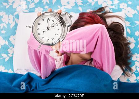 Junge Frau, die im Bett liegt und mit ihrem Arm ihre Augen bedeckt und einen Wecker hält. Student will nicht früh aufwachen für Universität oder Schule. Übergänge