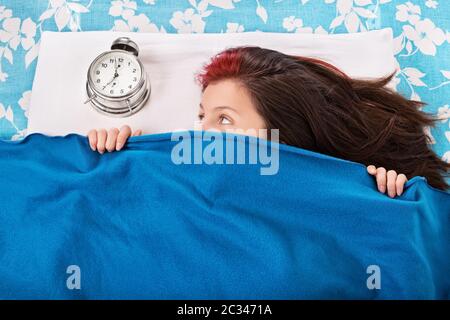 Porträt einer jungen Frau, die im Bett liegt, sich unter einer blauen Decke versteckt und auf den Wecker schaut. Student will nicht früh aufwachen für universi Stockfoto