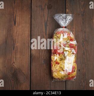 Transparente Zellophan Beutel mit Raw bunte Nudeln in Form von Bögen auf braunem Holz- Oberfläche, kopieren Raum Stockfoto