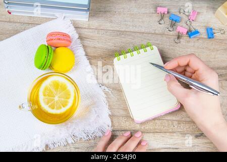 Die Hand hält einen silbernen Stift und schreibt in einem offenen Notizbuch, neben einem Notizbuch auf einem weißen Handtuch gibt es Minztee mit Zitronen- und mehrfarbigen Muffins Stockfoto