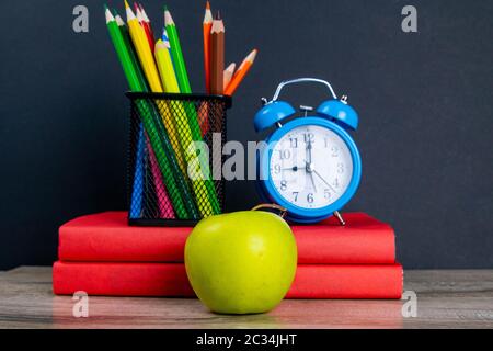 Der blaue Wecker mit mehrfarbigen Bleistiften steht auf einem Stapel roter Bücher neben einem Apfel. Stockfoto