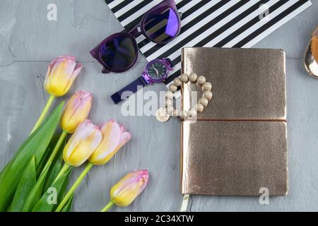 Ein goldenes Notizbuch mit Damenschuhen, Tulpen, Brillen und einer Uhr auf betonierten Hintergrund. Flet-Lay. Stockfoto