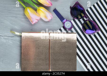 Ein goldenes Notizbuch mit Damenschuhen, Tulpen, Brillen und einer Uhr auf betonierten Hintergrund. Flet-Lay. Stockfoto