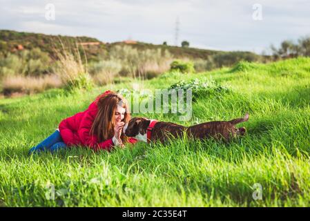 Junge Frau mit rotem Mantel und Jeans, die mit American Staffordshire Terrier auf dem Feld spielt Stockfoto