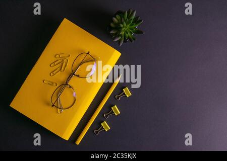 Gelber Bleistift, gelbes Notizbuch und Brille, Stifte, Clip-Papier auf schwarzem Hintergrund. Stockfoto