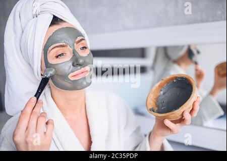 Schöne lächelnde Frau zu Hause im Bademantel mit einem Handtuch Anwendung Gesicht Tonmaske gegen Akne, um Problem Haut verjüngen Stockfoto