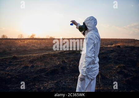 Laborarbeiter in weißem Overall, Gasmaske und Handschuhe beobachten Reagenzglas mit blauer Flüssigkeit während der Untersuchung verbranntes Gras und Erde auf verbranntem Gebiet bei untergehenden Sonne, Konzept der ökologischen Katastrophe Stockfoto