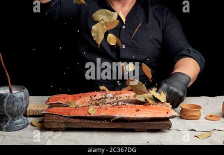 Erwachsener Mann in einem schwarzen T-Shirt gießt weißen groben Salz und einem trockenen Lorbeerblatt auf einem frischen Lachsfilet, Kochen, dunklen Hintergrund Stockfoto