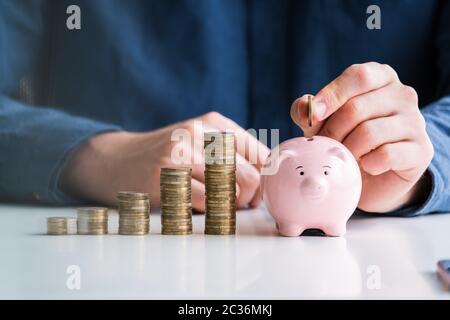 Person's Hand einfügen Münze in das Sparschwein mit zunehmender Münzen Stapel auf dem Schreibtisch Stockfoto