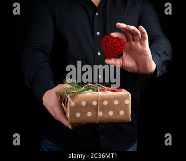 Erwachsener Mann steht auf einem dunklen Hintergrund, der ein schwarzes Shirt und eine verpackte Geschenk in braunem Papier in der Hand, und eine rote geschnitzten dekorative Er Stockfoto