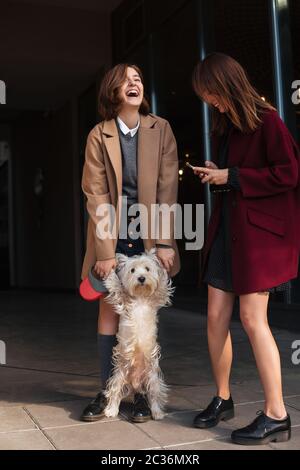 Portrait von zwei schönen lächelnden Mädchen stehen und glücklich spielen mit niedlichen kleinen Hund auf der Straße