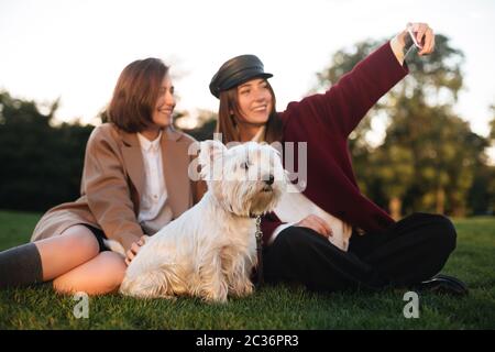Portrait von zwei jungen schönen Damen auf Gras sitzen und Selfie auf Handy, während die Zeit mit ihrem niedlichen Hund im Park Stockfoto
