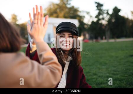 Nahaufnahme Foto von hübschen Dame auf Gras sitzen und geben fünf an ihre Freundin, während glücklich Zeit im Park verbringen Stockfoto