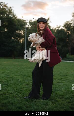 Hübsche lächelnde Dame in schwarzer Mütze und Mantel, die im Park steht und freudig in der Kamera schaut, während sie ihren kleinen niedlichen Hund umarmt Stockfoto