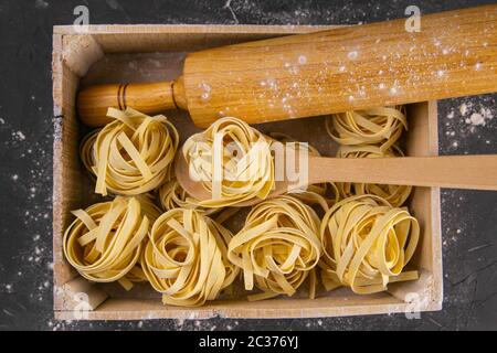 Stillleben der Zubereitung von Pasta auf rustikalem Holzhintergrund. Rohe Tagliatelle in einer Holzkiste mit Nudelholz für Teig und Holzlöffel. Top-Ansichten clos Stockfoto