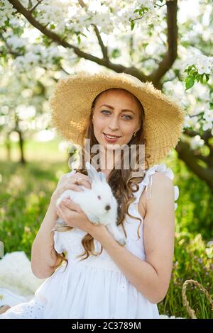 Das schöne Mädchen mit weißem Kaninchen im blühenden Apfelgarten. Weiße Blumen in einem Garten bei Sonnenschein. Frühling Apfelbäume in Blüte. Sehr Stockfoto