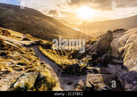 Fußweg und großer Felsbrocken, die ins Tal führen, mit Wasserreservoir, dramatischer Sonnenuntergang mit Sonnenstrahlen. Mou Stockfoto