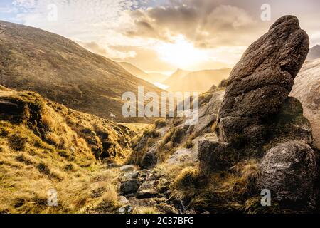 Großer Felsblock und Tal mit Wasserreservoir, dramatischer Sonnenuntergang mit Sonnenstrahlen. Mourne Mountains Stockfoto