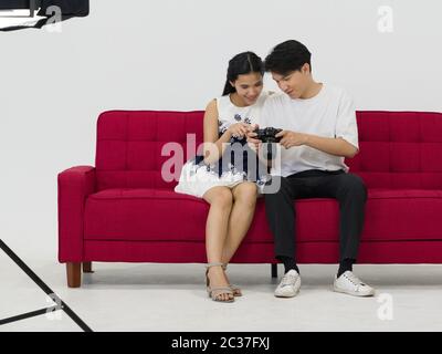 Asiatische Fotografen ermöglichen es Modellen, Bilder auf dem Kamerabildschirm anzusehen, während sie auf einem roten Sofa sitzen. Arbeitsatmosphäre im Fotostudio Stockfoto