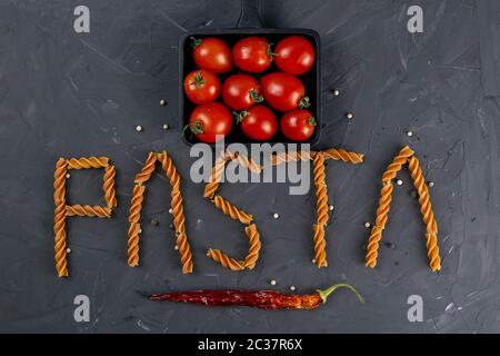Italienische Fusilli - Wort Pasta. Rohe italienische Pasta Fusilli mit Kirschtomaten in der Pfanne und heissem Papier auf dunklem Hintergrund von oben. Flaches Lay. Stockfoto