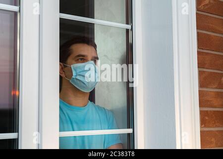 Junger Mann in medizinischer Maske schaut aus dem Fenster. Coronavirus-Pandemie. Home Quarantäne, Selbstisolierung wegen der Coronavirus-Krankheit, COVID-19. M Stockfoto