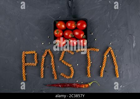 Italienische Fusilli - Wort Pasta. Rohe italienische Pasta Fusilli mit Kirschtomaten in der Pfanne und heissem Papier auf dunklem Hintergrund von oben. Flaches Lay. Stockfoto