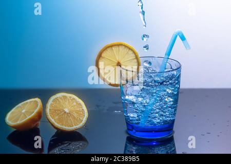 Ein Becher mit Eis und Wasser sitzt auf einem gespiegelten Tisch mit geschnittenen Zitronen. Zubereitung eines frischen Getränks Tonic, Saft oder Cocktail.Wasserspritzer,Wassertropfen.Splash Stockfoto