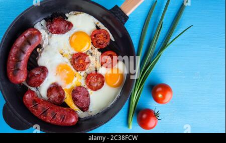 Bratwürste mit Eiern in gusseiserner Pfanne. Blauer Hintergrund. Blick von oben. Das Konzept eines traditionellen Mittagessens. Stockfoto