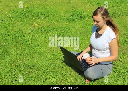 Ein schönes junges weißes Mädchen in einem weißen T-Shirt und mit langen Haaren auf grünem Gras sitzen, auf dem Rasen und arbeiten hinter einem schwarzen Laptop und halten eine Papiertasse mit Kaffee. Stockfoto