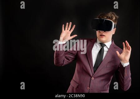 Jurastudent mit blonden Haaren in burgunderrot Jacke, weißes Hemd und schwarze Krawatte tragen Virtual Reality Brille posiert auf isoliertem schwarzen Hintergrund gekleidet Stockfoto