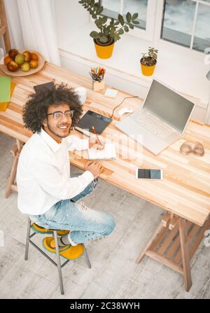 Kreativer Millennial-Mann, der am Arbeitsplatz zu Hause arbeitet. Junger arabischer Geschäftsmann schaut auf Kamera sitzen am Holzschreibtisch mit Computer, Handy und Stockfoto